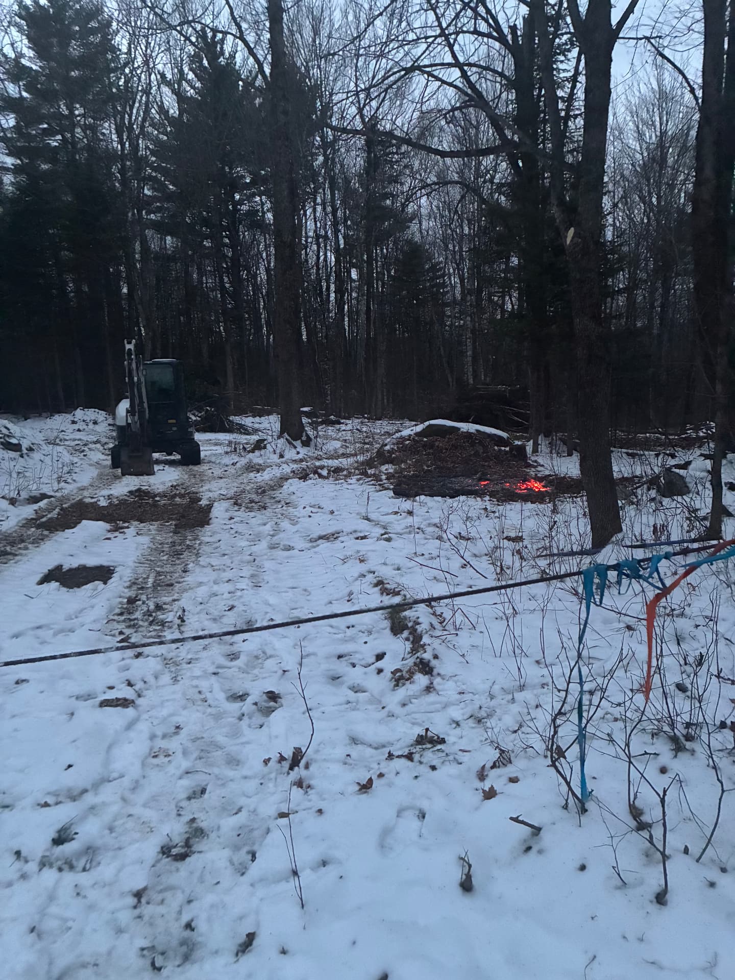 Mini excavator clearing a wooded lot in the snow with a brush pile burning